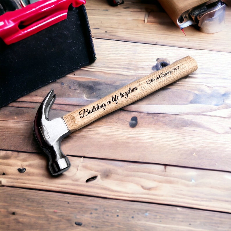 Op de afbeelding: Een houten hamer met een metalen kop, gegraveerd met de tekst "Building a life together" en de namen "Cella en Sydney" en het jaar "2022".