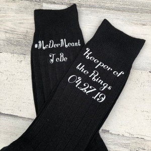 May include: A pair of black socks with white embroidered text. One sock reads "#McDerMeantToBe" and the other reads "Keeper of the Rings 04.27.19".