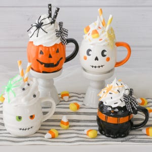 Puede incluir: Cuatro tazas tem&aacute;ticas de Halloween con coberturas de crema batida y pajitas decorativas. Las tazas tienen forma de calabaza, calavera, momia y gato negro. Caramelos de ma&iacute;z est&aacute;n esparcidos alrededor de las tazas.