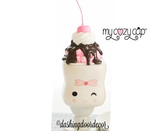 May include: A white ceramic mug shaped like an ice cream sundae with a pink cherry on top, chocolate sauce, and whipped cream. The mug has a cute cartoon face with a pink bow and a wink. The text "my cozy cup" is in the upper right corner of the image.