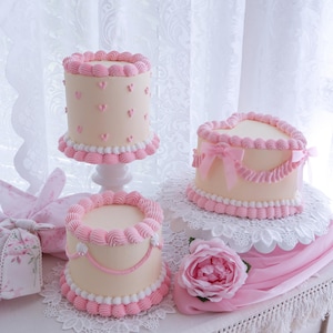 May include: Three decorative cakes with pink frosting and heart-shaped accents. One cake is heart-shaped with a pink bow. The cakes are on white pedestals and a lace doily. A pink rose and a wrapped gift are also present.