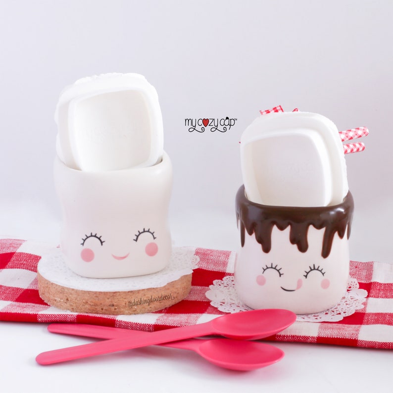 Puede incluir: Dos tazas de cer&aacute;mica blanca con caras sonrientes, una con un chorrito de chocolate y una tapa de malvavisco blanco, la otra con una tapa de malvavisco blanco liso. Las tazas est&aacute;n sobre un mantel a cuadros rojo y blanco con cucharas rosas.