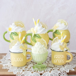 May include: A display of lemon-shaped mugs and a small glass bowl, all filled with whipped cream. The yellow mugs have green handles and are decorated with lemon slices, flowers, and straws. One mug says "SWEET" and another says "FRESH LEMONS".