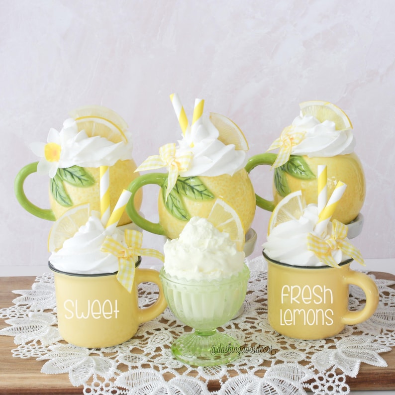 May include: A display of lemon-shaped mugs and a small glass bowl, all filled with whipped cream. The yellow mugs have green handles and are decorated with lemon slices, flowers, and straws. One mug says "SWEET" and another says "FRESH LEMONS".