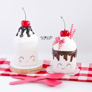 Puede incluir: Dos tazas de cer&aacute;mica blanca con caras sonrientes, decoradas para parecerse a helados. Una taza tiene salsa de chocolate y una cereza roja en la parte superior, la otra tiene crema batida y una cereza roja. Ambas tazas est&aacute;n sobre un mantel a cuadros rojo y blanco. "my cozy cup" est&aacute; escrito en el mantel.