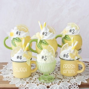 May include: A display of six lemon-themed decorative items. Features yellow mugs and a green glass, all adorned with white whipped cream, lemon slices, and straws. The mugs are inscribed with the words "Sweet" and "Fresh Lemons".