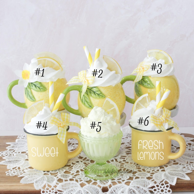 May include: A display of six lemon-themed decorative items. Features yellow mugs and a green glass, all adorned with white whipped cream, lemon slices, and straws. The mugs are inscribed with the words "Sweet" and "Fresh Lemons".