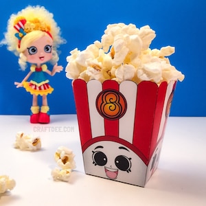May include: A red and white striped popcorn box with a cartoon face and the number 8 on the front. The box is filled with popcorn and is next to a doll with blonde hair and a blue and red dress.