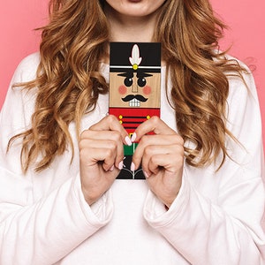 Puede incluir: Una mujer sostiene un adorno de cascanueces de madera con un uniforme rojo y verde, un sombrero negro con una pluma blanca y un bigote. El cascanueces tiene una cara pintada con ojos negros, un bigote negro y mejillas rojas.