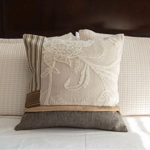 May include: A decorative pillow with a floral design and a striped accent, resting on a bed with white waffle-weave pillows. The pillow has a neutral color palette with brown and beige tones.