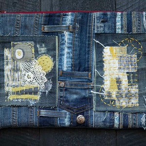 Pode incluir: Uma clutch retangular de ganga com zíper vermelho, apresentando designs de patchwork. Os patches incluem detalhes bordados em amarelo e branco, um pequeno bolso e um botão. A estética geral é uma mistura de estilos vintage e artesanais.