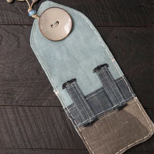 May include: A light blue fabric pencil case with a white button and three pockets. The case is made of a patchwork of different fabrics.