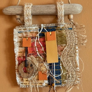 Può includere: Un piccolo arazzo da parete realizzato in juta, ritagli di tessuto e pelle. L'arazzo è attaccato a un pezzo di legno di deriva con uno spago. L'arazzo presenta una varietà di trame e colori, tra cui marrone, beige, arancione e blu.