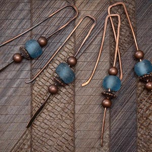 Pode incluir: Brincos de cobre artesanais com um design único. Cada brinco apresenta uma conta de vidro azul, contas de cobre e um elemento decorativo. Em um fundo de madeira texturizada.