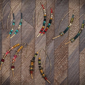 May include: A collection of handmade earrings with a variety of colorful beads and gold-tone wire hooks. The earrings feature turquoise, red, yellow, and green beads, creating a bohemian style. The earrings are displayed on a wooden surface.