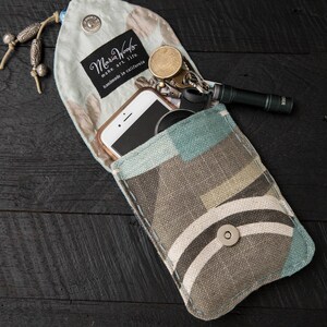 May include: A blue and brown fabric pouch with a snap closure. The pouch is open and contains a smartphone, keys, and a small flashlight. The pouch has a label that reads "Maria Woods make art life handmade in California."