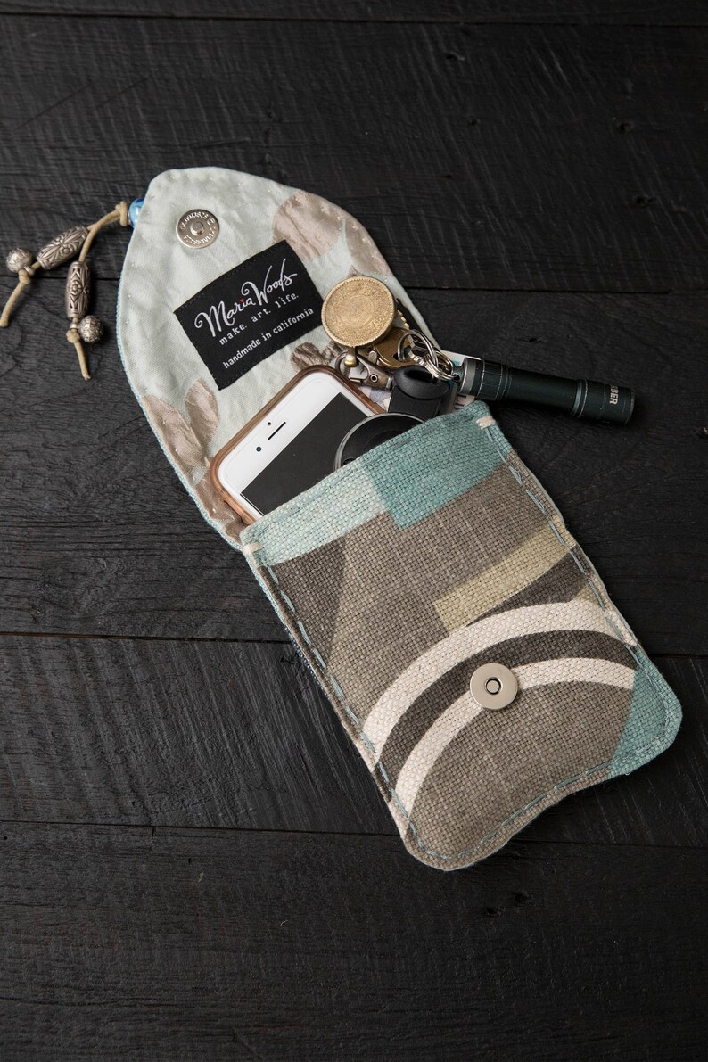 May include: A blue and brown fabric pouch with a snap closure. The pouch is open and contains a smartphone, keys, and a small flashlight. The pouch has a label that reads "Maria Woods make art life handmade in California."