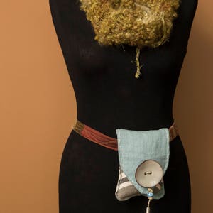 May include: A black dress form with a green and brown crocheted scarf around the neck. A brown and red belt with a light blue fabric pouch with a white button is attached to the dress form.