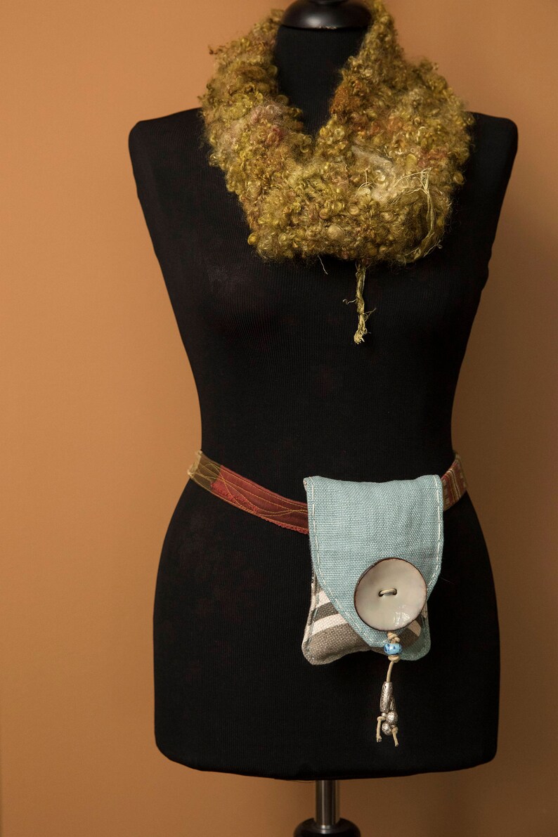 May include: A black dress form with a green and brown crocheted scarf around the neck. A brown and red belt with a light blue fabric pouch with a white button is attached to the dress form.