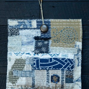 May include: A small, rectangular patchwork bag with a blue, white, and brown color scheme. The bag is made of different fabrics and has a variety of stitching patterns. A wooden bead necklace hangs from the top of the bag.