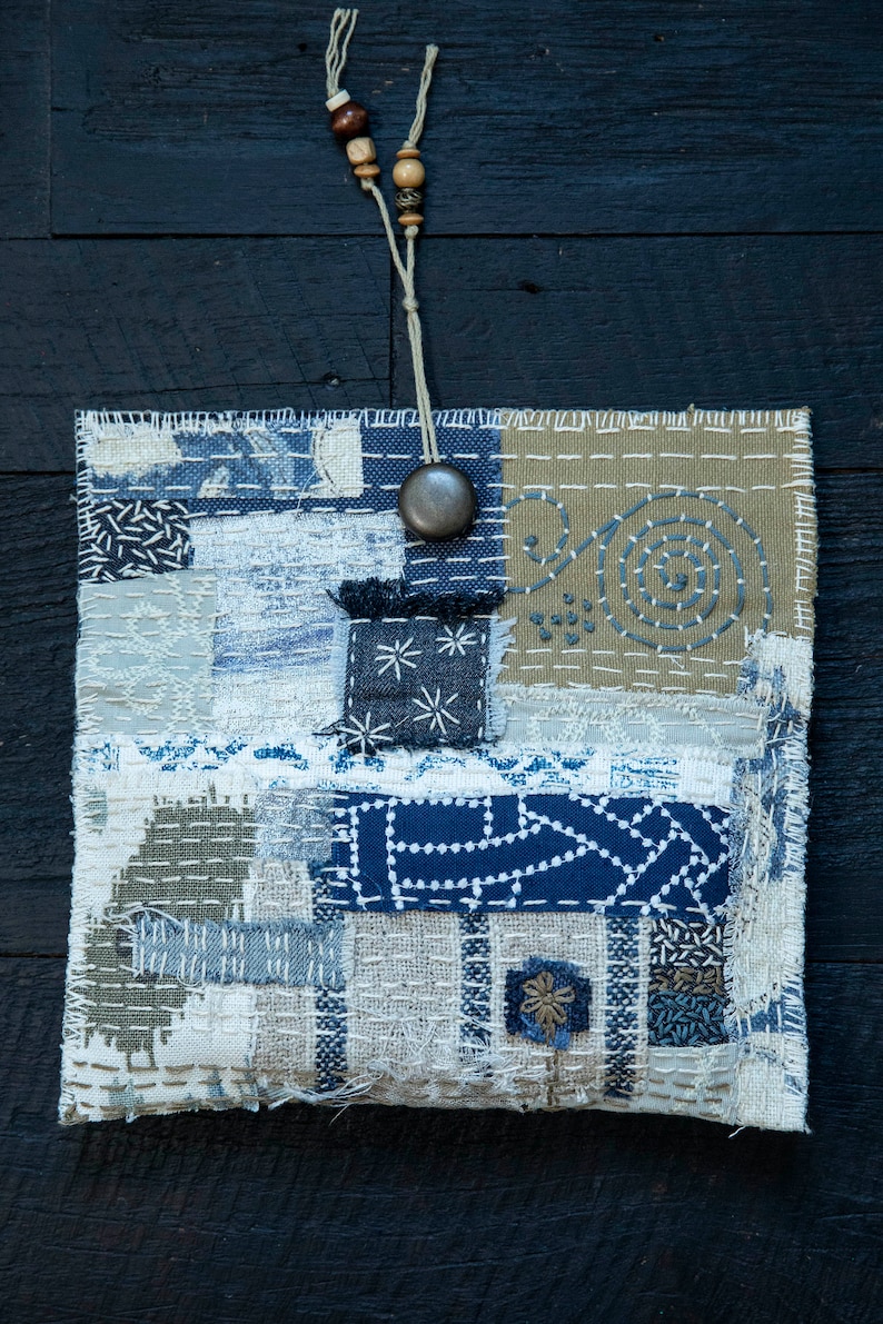 May include: A small, rectangular patchwork bag with a blue, white, and brown color scheme. The bag is made of different fabrics and has a variety of stitching patterns. A wooden bead necklace hangs from the top of the bag.