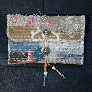 May include: A small, handmade patchwork pouch with a flap closure and two metal buttons. The pouch is decorated with a variety of fabrics, including denim, lace, and burlap. It has a beaded tassel hanging from the bottom.