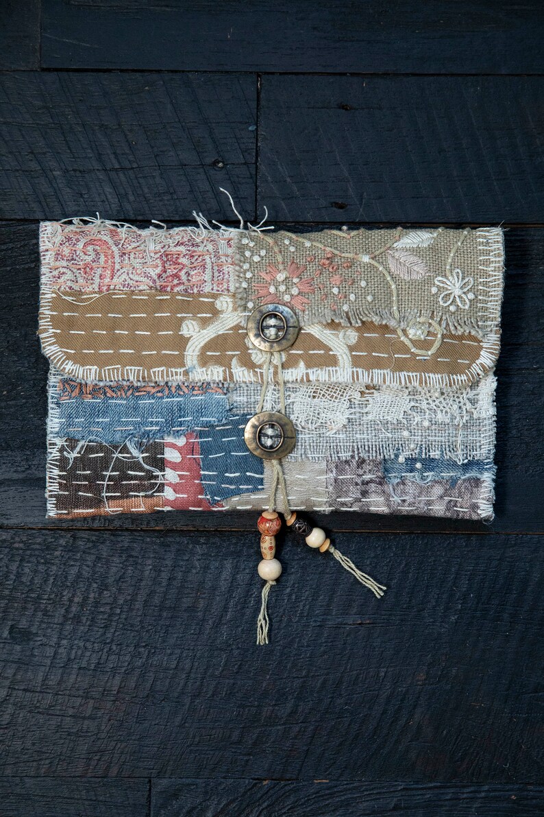 May include: A small, handmade patchwork pouch with a flap closure and two metal buttons. The pouch is decorated with a variety of fabrics, including denim, lace, and burlap. It has a beaded tassel hanging from the bottom.