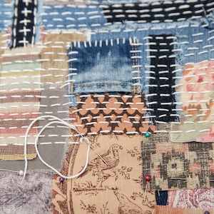 May include: A close-up of a patchwork quilt with various fabrics and textures. The quilt is made of different colors, including brown, blue, and white. The quilt is stitched together with white thread. There are several pins holding the fabric together.