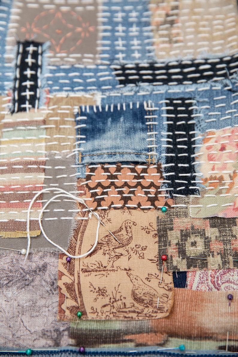 May include: A close-up of a patchwork quilt with various fabrics and textures. The quilt is made of different colors, including brown, blue, and white. The quilt is stitched together with white thread. There are several pins holding the fabric together.