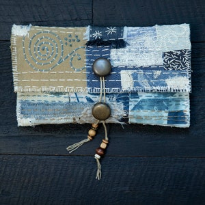 May include: A small, handmade patchwork pouch with a blue and beige color scheme. The pouch has two metal buttons and a beaded tassel.