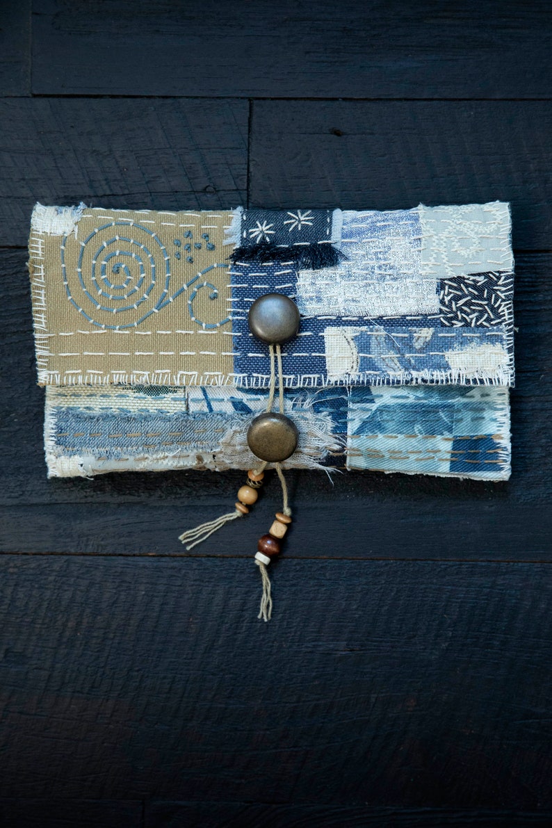 May include: A small, handmade patchwork pouch with a blue and beige color scheme. The pouch has two metal buttons and a beaded tassel.
