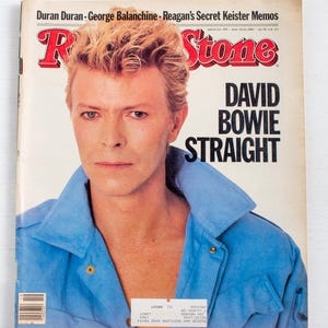 Peut inclure: Une couverture vintage du magazine Rolling Stone avec David Bowie. La couverture présente un titre rouge et blanc et le texte "DAVID BOWIE STRAIGHT". Bowie porte une veste bleue et a les cheveux blonds.
