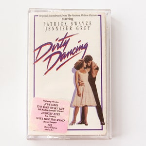 May include: A cassette tape case with a white background and a photo of Patrick Swayze and Jennifer Grey from the movie "Dirty Dancing". The text "Dirty Dancing" is in purple and blue lettering. The case also features a sticker with the text "I've Had The Time Of My Life", "Hungry Eyes", "She's Like The Wind", and "Yes".