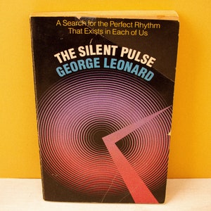 May include: A black book cover with a colourful circular design and the text "THE SILENT PULSE GEORGE LEONARD" in white and blue letters. A Search for the Perfect Rhythm That Exists in Each of Us.