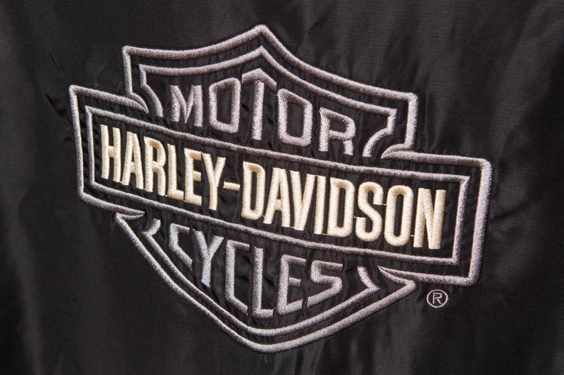 May include: A black embroidered patch with the Harley-Davidson logo in white and gray. The logo features the words "Motor", "Harley-Davidson", and "Cycles" in a shield shape.