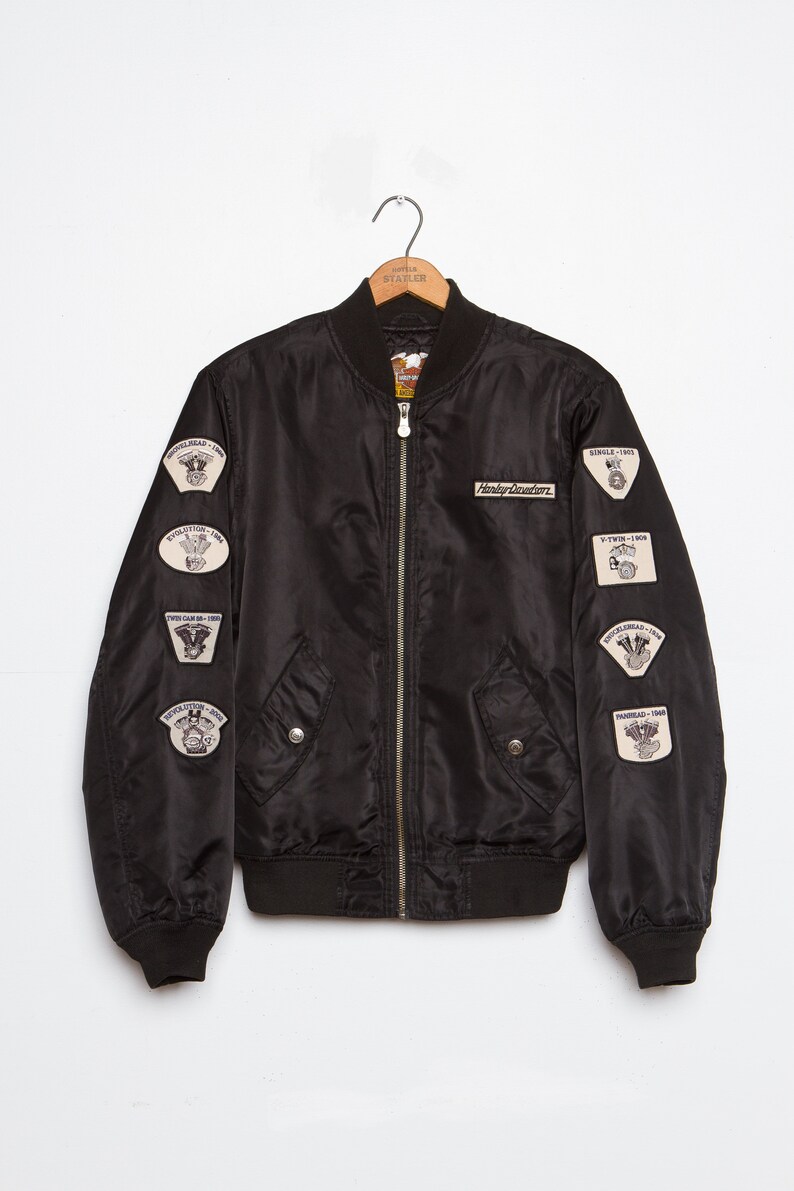 May include: Black bomber jacket with multiple Harley Davidson patches on the sleeves. The patches feature different motorcycle engine designs and dates.