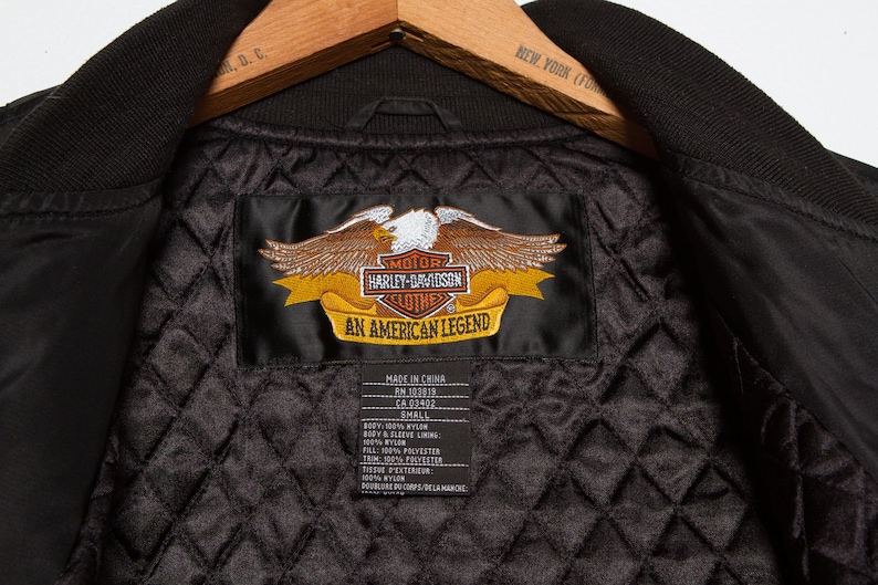 May include: Black Harley-Davidson jacket with a quilted lining and a satin label that reads "Motor Harley-Davidson Clothes An American Legend". The label has an embroidered eagle with a yellow banner.
