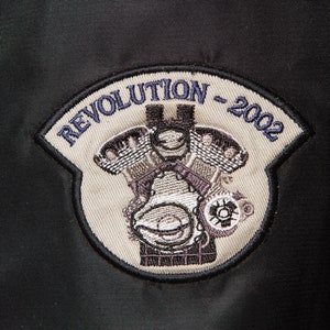 May include: A beige embroidered patch with the text "Revolution - 2002" in blue thread. The patch features a detailed image of a motorcycle engine.
