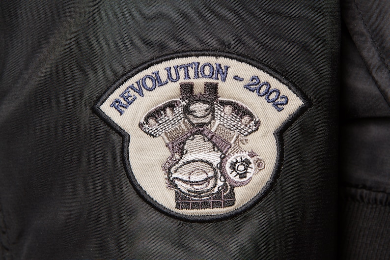 May include: A beige embroidered patch with the text "Revolution - 2002" in blue thread. The patch features a detailed image of a motorcycle engine.