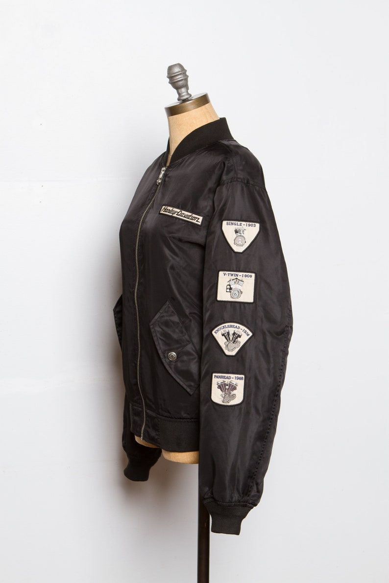 May include: Black bomber jacket with a Harley Davidson logo and patches on the sleeve. The patches feature the text "Single - 1903", "V-Twin - 1909", "Knucklehead - 1936", and "Panhead - 1940".