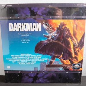 May include: A VHS tape cover for the film "Darkman", featuring a shadowy figure in a long coat and hat standing in front of a fiery backdrop. The text "Darkman" is in large, bold letters at the top of the cover.