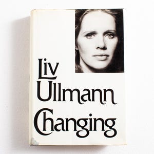 以下が含まれることがあります： リヴ・ウルマン著「チェンジング」のヴィンテージ本の表紙。表紙は白で、黒い文字と女性の白黒ポートレートが描かれています。本は少し擦り切れています。