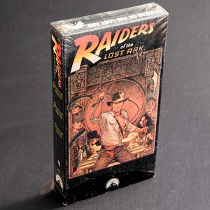 May include: A vintage VHS tape case for the movie Raiders of the Lost Ark. The case features a colorful illustration of Indiana Jones holding a whip and surrounded by other characters from the film.