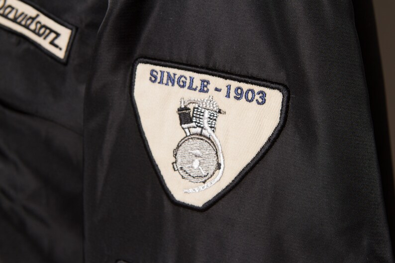 May include: A black patch with white stitching and the text "SINGLE - 1903" in blue. The patch features a black and white illustration of a motorcycle engine.