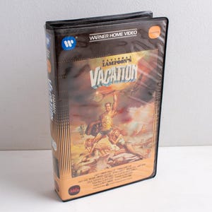Op de afbeelding: Een zwarte plastic VHS-hoes voor de film National Lampoon's Vacation. De cover bevat een kleurrijke illustratie van een persoon die een tennisracket boven het hoofd houdt, met de filmtitel in witte tekst en Warner Home Video-branding.