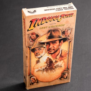 May include: A VHS tape case for the movie Indiana Jones and the Last Crusade. The cover features a close-up of Indiana Jones, wearing his iconic fedora, and his father, Henry Jones Jr., with the movie title in bold letters.