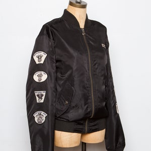 May include: A black bomber jacket with a zipper closure and multiple patches on the left sleeve. The patches feature motorcycle engine designs and the text "Shovelhead - 1969", "Evolution - 1984", "Twin Cam - 1999", and "Revolution - 2002".