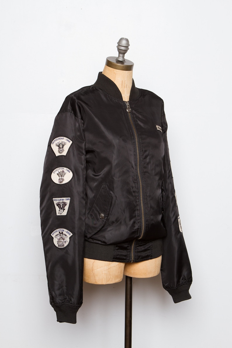 May include: A black bomber jacket with a zipper closure and multiple patches on the left sleeve. The patches feature motorcycle engine designs and the text "Shovelhead - 1969", "Evolution - 1984", "Twin Cam - 1999", and "Revolution - 2002".