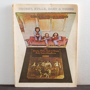 Könnte beinhalten: Ein Vintage-Vinyl-Plattencover mit einem Foto von Crosby, Stills & Nash. Das Cover ist hellbraun mit einem Schwarzweißfoto der Band, die auf einem Sofa sitzt. Der Albumtitel "Crosby, Stills, Nash & Young" ist in weißen Buchstaben oben auf dem Cover gedruckt. Der Albumtitel "So Far" ist in weißen Buchstaben unten auf dem Cover gedruckt.
