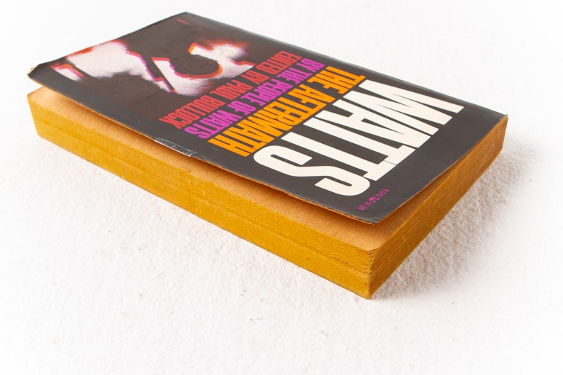 May include: A paperback book titled "The Aftermath" by the author "Watts" with the text "VICTOR GOLLANCZ LTD" on the bottom right corner of the cover. The cover is black with pink, orange, and yellow text.